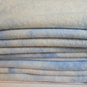 May include: A stack of folded fabric featuring a soft, fuzzy texture with a mottled pattern of light blue and white. The material appears thick and plush, with multiple layers neatly piled on top of each other, showing a consistent colour blend.