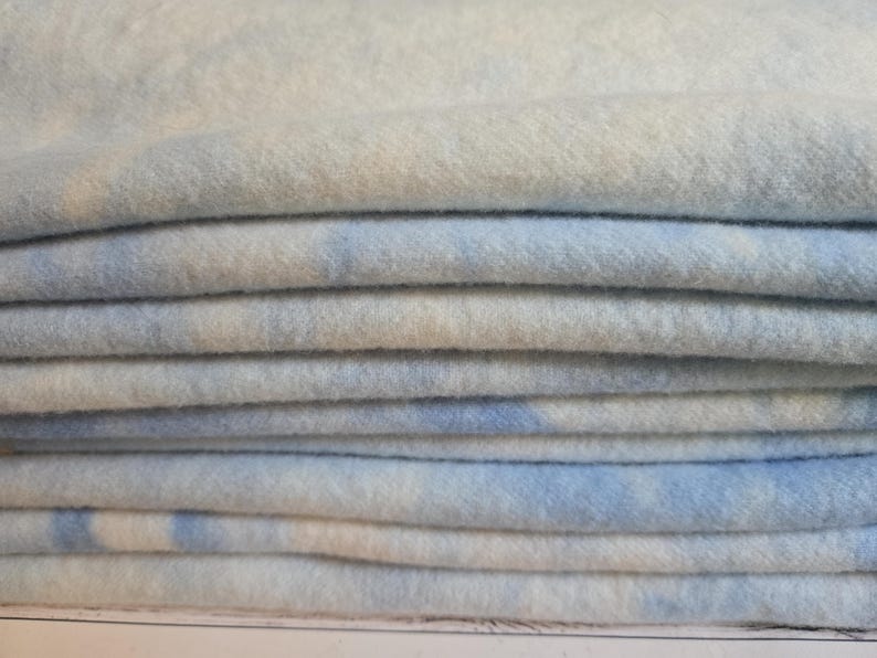 May include: A stack of folded fabric featuring a soft, fuzzy texture with a mottled pattern of light blue and white. The material appears thick and plush, with multiple layers neatly piled on top of each other, showing a consistent colour blend.