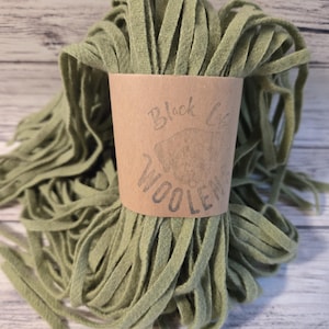 May include: A bundle of sage green wool strips, wrapped with a brown paper label. The label reads "Black Cat Woolens" in a handwritten style. The wool strips are arranged in a circular shape, with the ends of the strips visible.
