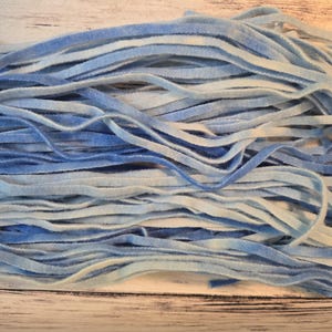 May include: A collection of thin, flat velvet ribbons in varying shades of blue and white. The ribbons feature a soft, textured surface with a gradient colour effect, transitioning between light sky blue, deep royal blue, and crisp white tones.