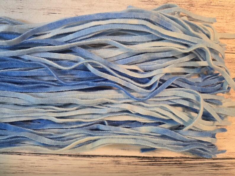 May include: A collection of thin, flat velvet ribbons in varying shades of blue and white. The ribbons feature a soft, textured surface with a gradient colour effect, transitioning between light sky blue, deep royal blue, and crisp white tones.