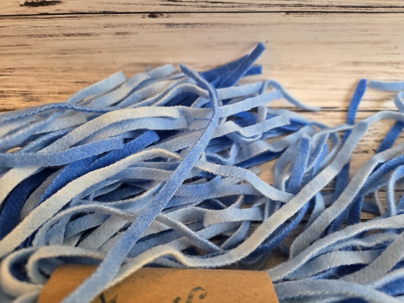 May include: A collection of thin, flexible strips in various shades of blue, ranging from light sky blue to deep royal blue. The strips have a soft, velvety texture and are bundled together, with a small portion of a brown paper tag visible at the base.