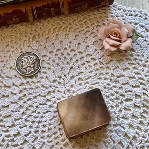 May include: A small rectangular box with a smooth, mottled brown and tan surface, a silver Mercury dime, and a pale pink ceramic rose flower. The box features a metallic trim along its edges, providing a contrast to the patterned top surface.