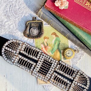 May include: A vintage rhinestone belt buckle with a black velvet band. The buckle is made of metal and has a geometric design with multiple rows of clear rhinestones. The belt is lying on a white surface with a vintage postcard and a small antique pocket watch.