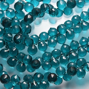 May include: A strand of teal blue faceted gemstone beads. The beads are round and have a pointed tip. The beads are strung on a clear thread.