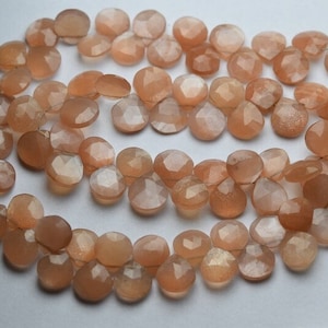 以下が含まれることがあります： ピーチ色のサンストーンビーズのネックレス。各ビーズはファセットカットされており、真珠のような光沢があります。ビーズは銀色のワイヤーに通されています。