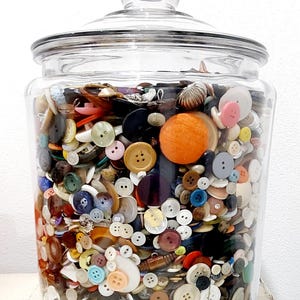 May include: A large clear glass jar filled with a colorful assortment of buttons. The buttons vary in size, shape, and color, including white, black, brown, blue, and orange. The jar has a glass lid.