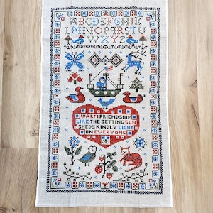 May include: A cross-stitch sampler with a cream-colored background. The design features an alphabet, various animals, a boat, and a heart with the text "WARM FRIENDSHIP LIKE THE SETTING SUN SHEDS KINDLY LIGHT ON EVERYONE". The border is a dark blue and red pattern.
