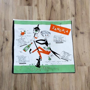 May include: A vintage Sangria recipe tea towel with a whimsical illustration. The design features a cartoon figure, a bottle of wine, and recipe instructions in multiple languages. The towel has a white background with green and black borders.