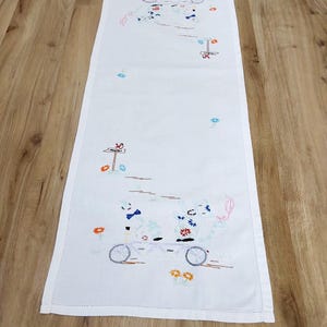 May include: White embroidered table runner featuring a tandem bicycle with two figures, flowers, and a signpost. The embroidery uses blue, orange, pink, and green thread. The runner is rectangular and has a stitched border.