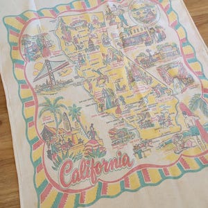 May include: Cream-colored kitchen towel featuring a colorful map of California with iconic landmarks and cities. The map is framed with a decorative border in pink, yellow, and teal. The word "California" is written in a cursive font.