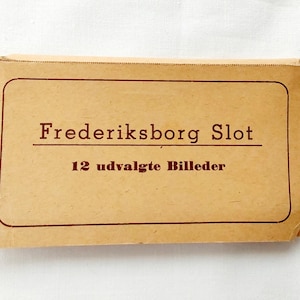May include: A vintage, rectangular card with rounded corners, featuring the text "Frederiksborg Slot" and "12 udvalgte Billeder" in a dark red font. The card has a light brown color and is set against a white background.