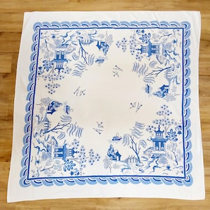 May include: White square tablecloth with a blue and white Asian-inspired design. The border features a wave pattern, and the center showcases a detailed landscape with pagodas, trees, and birds. The tablecloth is made of a soft, likely cotton material.