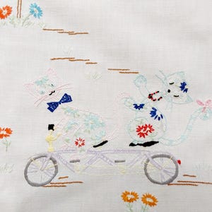 May include: Embroidered image of two cats riding a tandem bicycle. The cats are light blue and white with floral patterns and bow ties. The background features flowers and simple landscape details, all in soft colors.