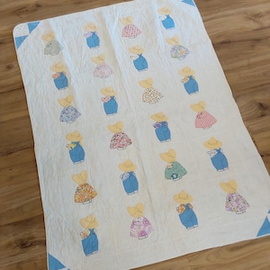 May include: A white rectangular quilt featuring a repeating pattern of appliqued figures wearing yellow sun hats and blue overalls. The figures are arranged in vertical rows, each wearing a different patterned shirt. The quilt has blue corner accents.