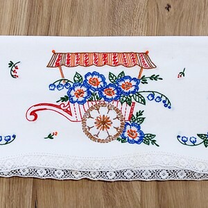 May include: White embroidered linen towel featuring a wheelbarrow filled with blue and orange flowers. The wheelbarrow has a red and white striped canopy. The towel has a white lace trim along the bottom edge.