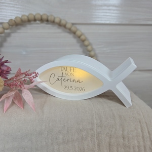 Könnte beinhalten: Eine weiße, fischförmige Dekorationsleuchte mit dem Text "TAUFE VON Caterina 29.5.2026" in einem goldfarbenen Oval. Das Licht liegt auf einer beigen Oberfläche, mit einem Blumenarrangement und Holzperlen im Hintergrund.