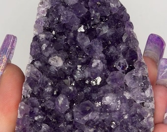 May include: A cluster of amethyst crystals, showcasing a deep purple hue with sparkling facets. The natural formation has a rough, irregular shape, with visible crystalline structures. The image highlights the stone's texture and color variations.
