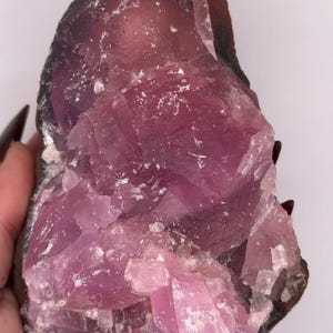 May include: A raw, uncut crystal formation with a striking blend of colors. The stone features a deep purple hue that transitions to a lighter pink, with areas of white. The rough edges and natural texture of the crystal are visible.