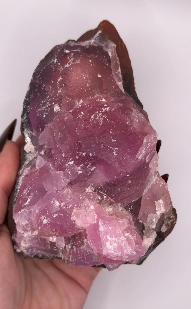 May include: A raw, uncut crystal formation with a striking blend of colors. The stone features a deep purple hue that transitions to a lighter pink, with areas of white. The rough edges and natural texture of the crystal are visible.