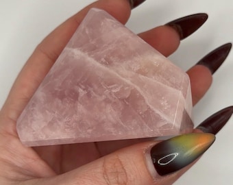 May include: A faceted rose quartz crystal shaped like a pyramid with a triangular base. The stone features a translucent, pale pink hue with visible internal fractures and natural mineral inclusions throughout its polished, geometric structure.