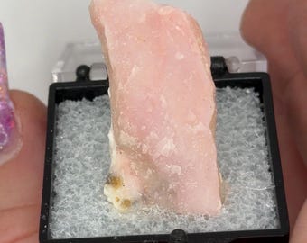 May include: A light pink opal gemstone displayed in a black plastic case with a clear lid. The opal has a rough, textured surface and a pale, translucent appearance. The case is lined with a gray, textured material.