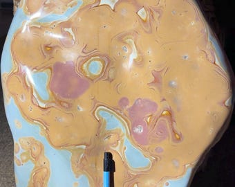 May include: Large, polished stone with a unique pattern of tan, light blue, and pink swirls. The stone has an irregular shape and a smooth surface. A blue pen is placed next to the stone for scale. This decorative stone could be used as a paperweight or display piece.