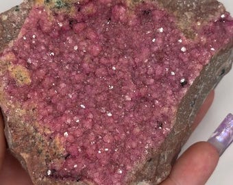May include: A raw mineral specimen featuring a central cluster of vibrant, deep pink, crystalline formations embedded within a rough, brownish-gray rock matrix. The surface of the pink crystals is textured with small, rounded, sparkling nodules.