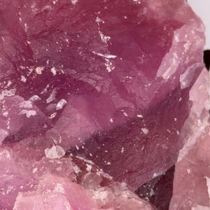 May include: Close-up of a rough, crystalline mineral with varying shades of pink and magenta. The surface has a textured appearance with visible facets and some white inclusions. The overall aesthetic suggests a natural, geological specimen.