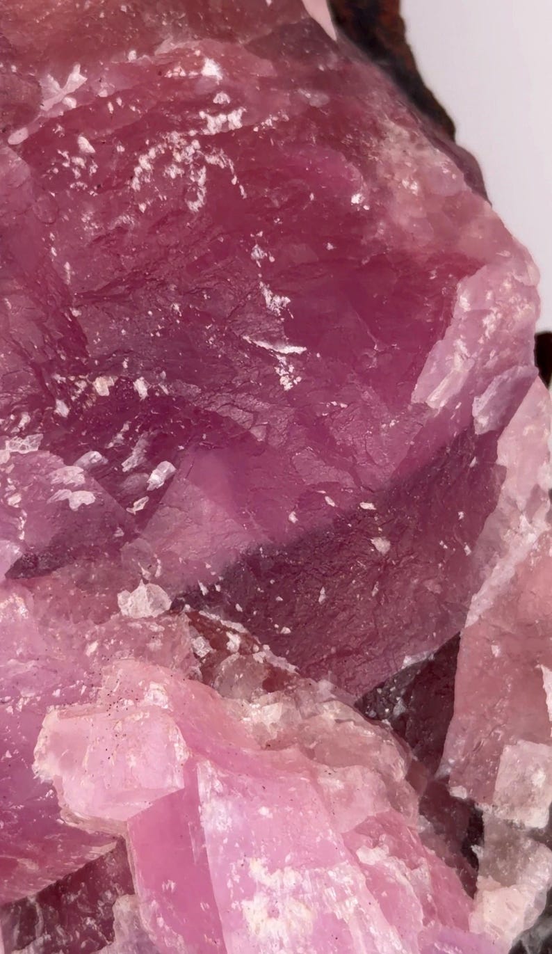 May include: Close-up of a rough, crystalline mineral with varying shades of pink and magenta. The surface has a textured appearance with visible facets and some white inclusions. The overall aesthetic suggests a natural, geological specimen.