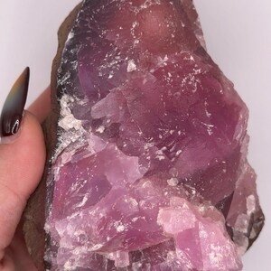 May include: A raw, uncut pink amethyst crystal. The stone has a deep purple hue at the edges, transitioning to a lighter pink in the center. The crystal's surface is rough and textured, with visible crystalline structures.