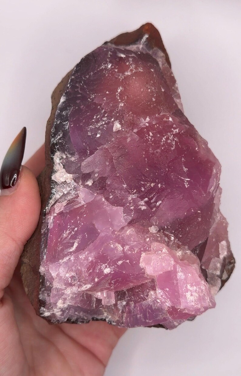 May include: A raw, uncut pink amethyst crystal. The stone has a deep purple hue at the edges, transitioning to a lighter pink in the center. The crystal's surface is rough and textured, with visible crystalline structures.