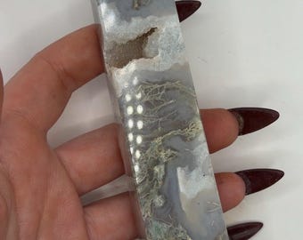 May include: A rectangular crystal obelisk with a pointed top. The stone features a mix of gray, white, and tan colors with intricate patterns. The crystal is held in a hand against a white background.