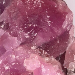 May include: Close-up of a rough, crystalline mineral specimen. The stone features shades of pink and purple, with a textured surface and visible crystal formations. The mineral has a dark brown edge.