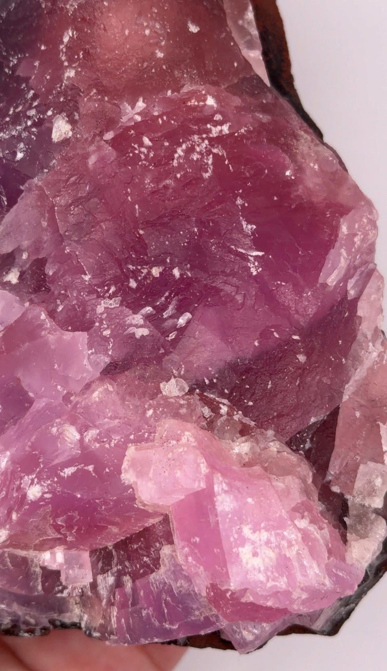 May include: Close-up of a rough, crystalline mineral specimen. The stone features shades of pink and purple, with a textured surface and visible crystal formations. The mineral has a dark brown edge.