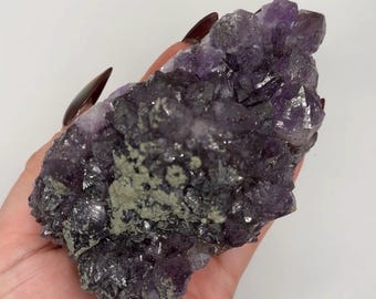 May include: A raw amethyst crystal cluster featuring deep purple, multifaceted points and a textured, grayish-green mineral inclusion on its surface. The cluster displays a jagged, natural formation with varying shades of violet and dark purple throughout.