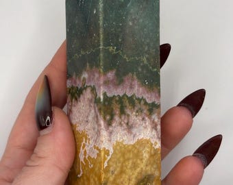 May include: A polished stone tower with a pointed top, featuring distinct horizontal bands of color. The top section is a deep forest green, transitioning into a thin layer of pink and white, with a textured, mustard yellow base at the bottom.