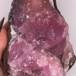 May include: A raw, uncut pink fluorite crystal with a dark brown matrix. The crystal has a layered appearance with a mix of light and dark pink hues. White flecks are scattered throughout the crystal.