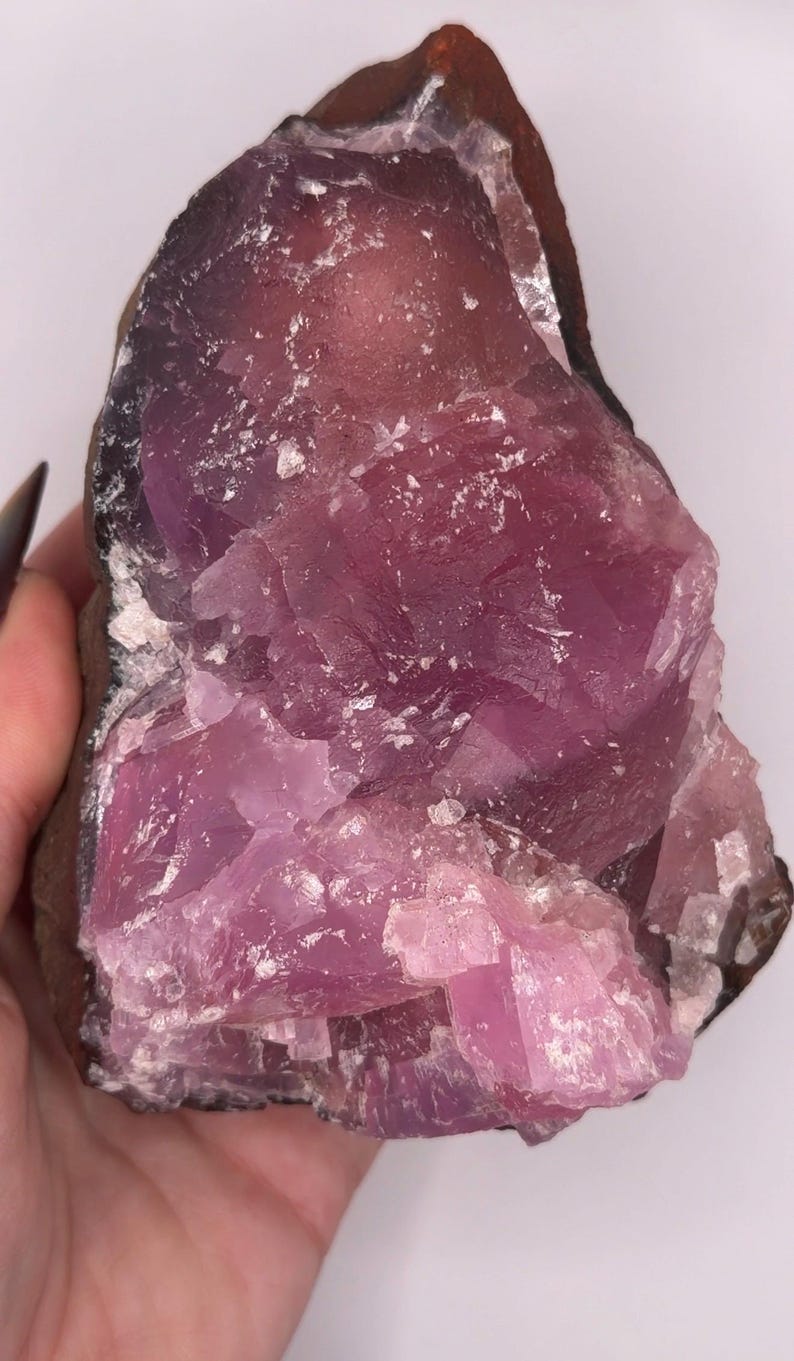 May include: A raw, uncut pink fluorite crystal with a dark brown matrix. The crystal has a layered appearance with a mix of light and dark pink hues. White flecks are scattered throughout the crystal.