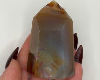 May include: A polished, faceted stone tower featuring translucent layers of gray, brown, and amber hues. The crystal point has a smooth, multi-sided surface with visible internal banding and natural color variations throughout the mineral structure.