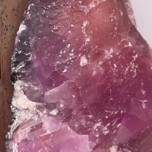 May include: Close-up of a raw, natural mineral specimen. The stone features a gradient of colors, from deep purple to light pink, with white crystalline inclusions. The rough texture and varied hues suggest a geological formation.