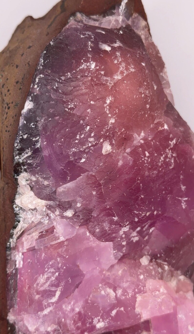 May include: Close-up of a raw, natural mineral specimen. The stone features a gradient of colors, from deep purple to light pink, with white crystalline inclusions. The rough texture and varied hues suggest a geological formation.