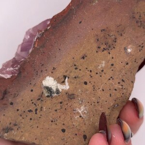 May include: A rough-hewn, reddish-brown stone with a textured surface and dark spots. A section of light purple crystal is visible on one edge. The stone is held in a hand.