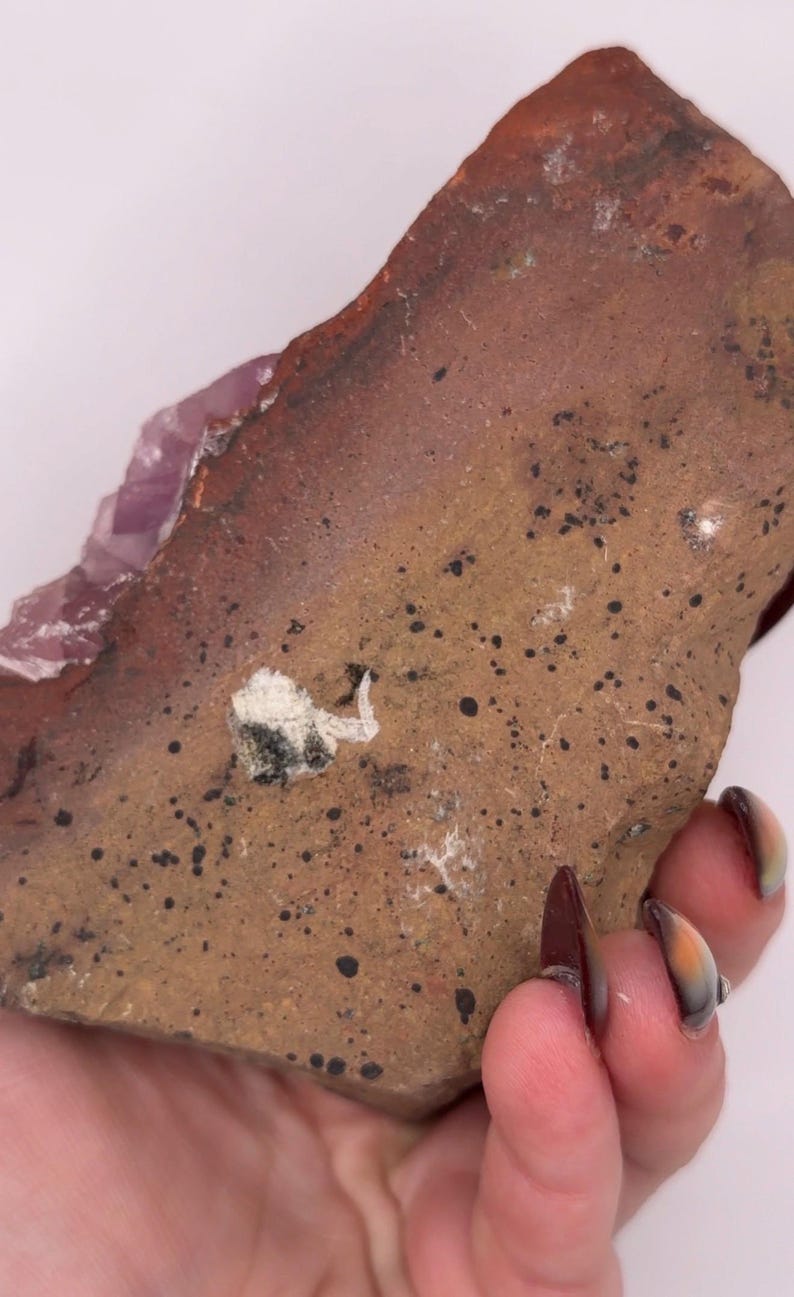 May include: A rough-hewn, reddish-brown stone with a textured surface and dark spots. A section of light purple crystal is visible on one edge. The stone is held in a hand.