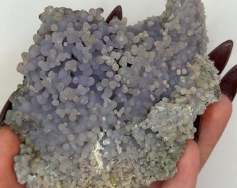May include: A piece of grape agate, a unique mineral formation. The surface is covered in small, round, light purple and gray spheres, resembling a cluster of grapes. The base has a rough, textured appearance with hints of green.