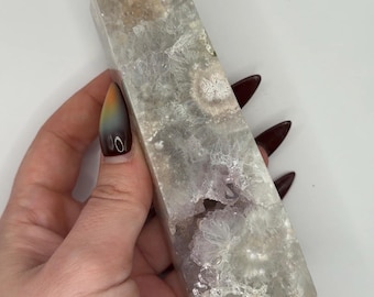 May include: A rectangular crystal tower with a polished, faceted surface. The crystal features a mix of translucent white, gray, and purple hues, with visible internal patterns. The crystal is held in a hand.