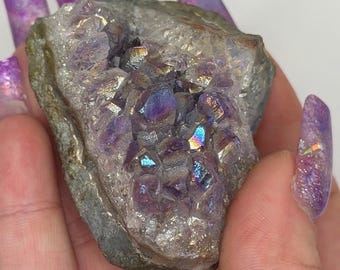 May include: A raw, natural amethyst cluster with iridescent, rainbow-like flashes. The crystal formation features a mix of deep purple and shimmering, metallic hues, set within a rough, gray matrix. The cluster is held in a hand, showcasing its natural beauty.