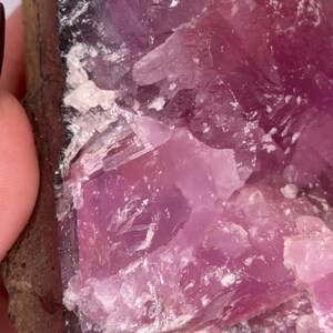 May include: Close-up of a rough, natural mineral specimen. The stone features a vibrant, translucent pink hue with crystalline structures and a dark brown matrix. The surface has a rough, uneven texture, with areas of white and light pink.