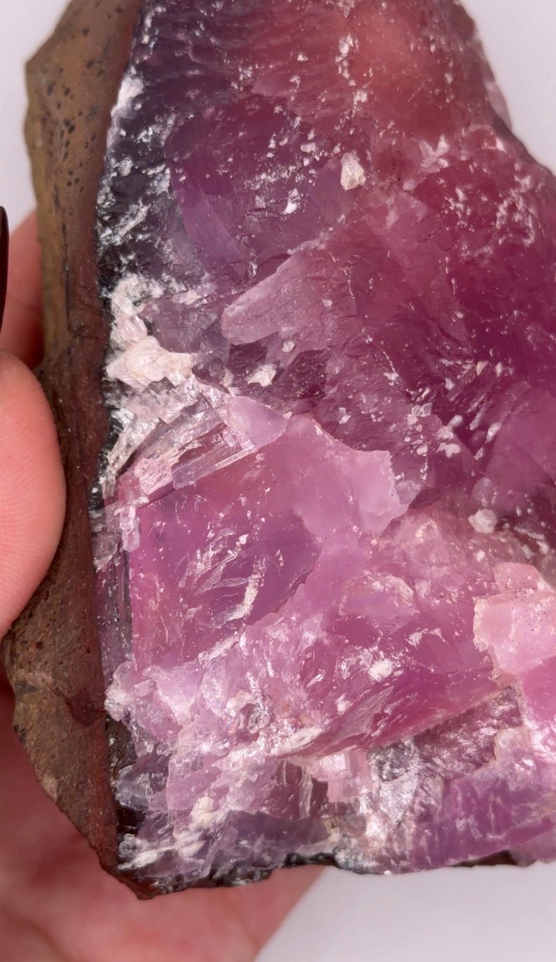 May include: Close-up of a rough, natural mineral specimen. The stone features a vibrant, translucent pink hue with crystalline structures and a dark brown matrix. The surface has a rough, uneven texture, with areas of white and light pink.