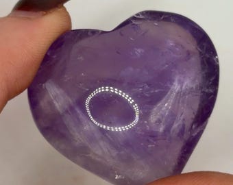May include: A polished, heart-shaped amethyst gemstone with a deep purple hue and translucent quality. The stone features natural internal inclusions and a smooth, rounded surface that reflects light, creating a bright circular highlight on its face.